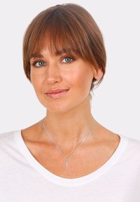 Strieborný náhrdelník s príveskom v tvare listu, nosený na holom krku, doplnený bielou košeľou a prirodzeným odtieňom pleti. Hladká metalická textura.
