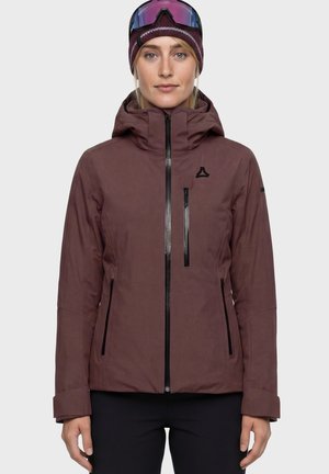 Frau trägt eine dunkelrote Winterjacke mit Kapuze und schwarzen Reißverschlüssen, eine dunkelrote Strickmütze und blau getönte Skibrille auf der Stirn.