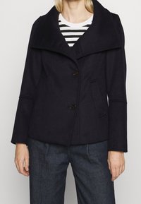 Abrigo de mezcla de lana azul marino con un collar ancho, dos botones grandes, bolsillos laterales y mangas acampanadas, llevado sobre una blusa de rayas.
