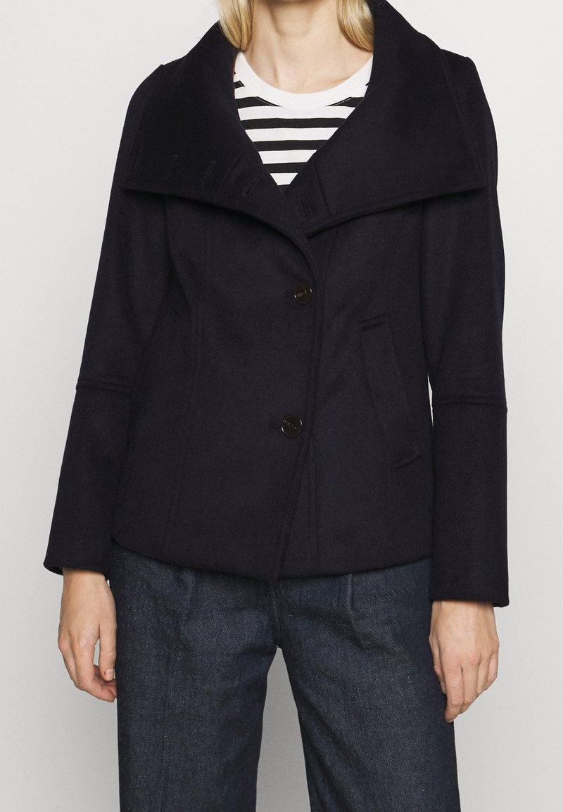 Abrigo de mezcla de lana azul marino con un collar ancho, dos botones grandes, bolsillos laterales y mangas acampanadas, llevado sobre una blusa de rayas.