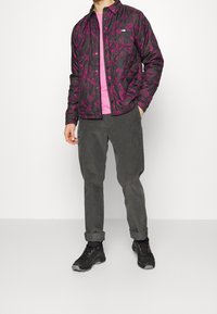 Vadderad, svart jacka med rosa blommönster, tillsammans med en rosa skjorta och grå manchesterbyxor. Svarta skor med röda detaljer.