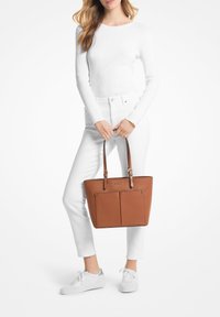 Bolso tote texturizado marrón con dos bolsillos frontales y asas de hombro marrones. La modelo lleva una blusa y pantalones blancos, sosteniendo el bolso. Fondo blanco.