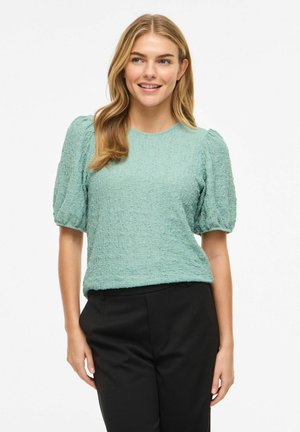 Uśmiechnięta kobieta z długimi blond włosami, ubrana w teksturowaną jasnooliwkową bluzkę z bufkami i czarne spodnie, stoi na białym tle.