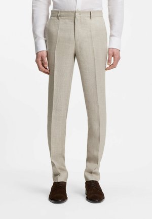 Beżowe, szyte na miarę spodnie lniane z przednimi plisami, noszone z ciemnobrązowymi eleganckimi butami i białą koszulą zapinaną na guziki.