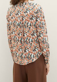 Blusa floreale bianca con motivo arancione e verde petrolio. Maniche lunghe con orlo arrotondato e dettaglio con piega sul retro. Tessuto leggero.