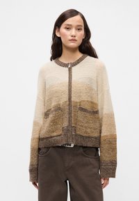 Πλεχτό γιλέκο με φερμουάρ μπροστά, με γήινες αποχρώσεις και οριζόντιες γραδίες. Το υλικό φαίνεται μαλακό και υφαντό με τσέπες.