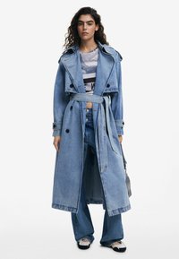 Denim-Trenchcoat in Hellblau, langes Design mit tailliertem Gürtel, ausgestattet mit doppelreihiger Knopfleiste und breiten Revers. Kombiniert mit passenden Jeans.