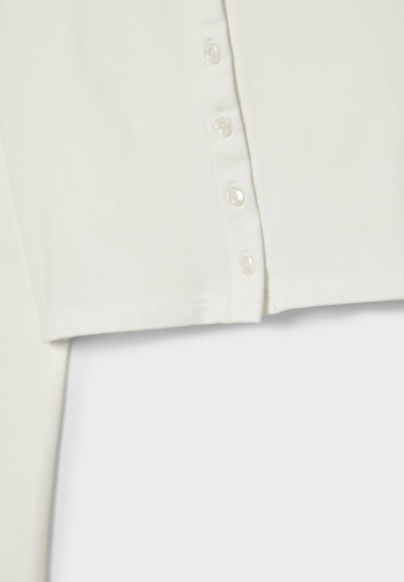 Top corto bianco realizzato in un tessuto morbido, con una silhouette dritta e sei bottoni perlacei che decorano la parte anteriore. Texture liscia e finitura pulita.