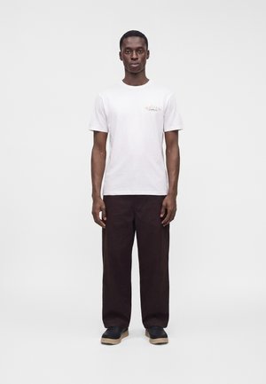 Homme debout portant un t-shirt blanc à manches courtes avec un petit imprimé sur la poitrine, un pantalon ample marron foncé et des chaussures noires avec des semelles beiges.