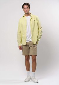 Junger Mann, der ein hellgelbes Hemd über einem weißen T-Shirt, beige Shorts, weiße Socken und weiße Sneaker trägt, vor einem neutralen Hintergrund steht.