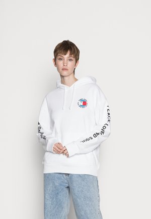 Hvit hettegenser med front kengurulomme, med et sirkulært logo og svart tekst langs ermene. Tøyet ser mykt og koselig ut.