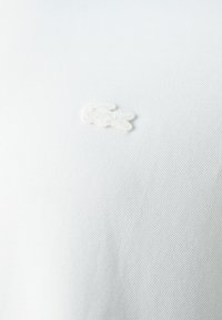 Tissu blanc texturé avec un logo de crocodile blanc brodé à la surface.