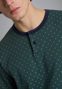 Camicia verde con motivo a fantasie caratterizzata da piccoli punti blu e rossi, colletto blu navy, due bottoni sul collo e un tessuto morbido e leggero.