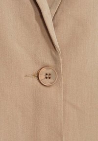 Beige tygblazer i nära bild, med en slät yta, en stor knapp med fyra hål och en notch-lapel-design.