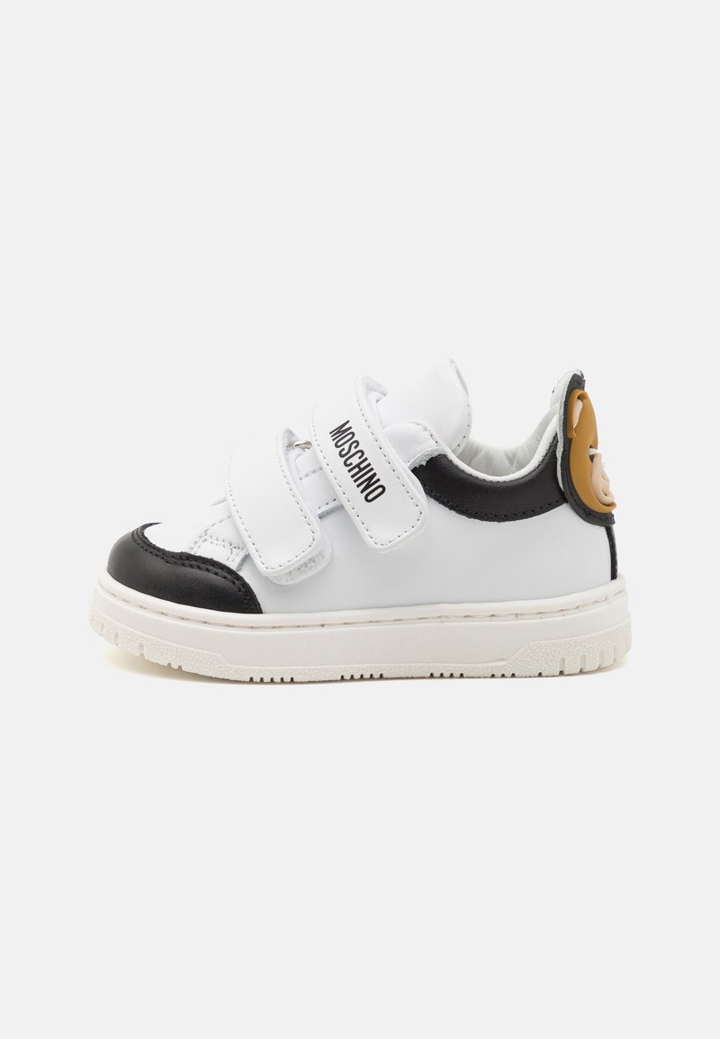 Zapatilla de cuero blanca y negra con dos correas de velcro, puntera redonda, suela texturizada y un detalle en marrón en el talón.