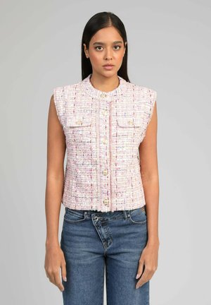 Jeune femme portant un gilet sans manches en tweed rose à boutons avec poches avant et jean taille haute bleu, debout devant un fond uni.