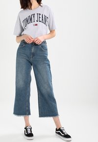 Camiseta gris con "TOMMY JEANS" impreso en azul marino, combinada con vaqueros de denim azul cortos con el dobladillo deshilachado y zapatillas negras.