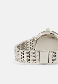 Silverfärgad metallklocka med en nätarmband, som har ett runt urtavla och en spänne. Ytan är polerad med inristad varumärkning på spännet.