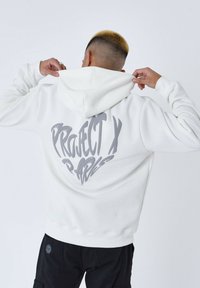 Sudadera blanca con capucha y un gráfico gris que dice "PROJECT X PARIS" en la parte trasera. Tejido suave, corte relajado, acabada con capucha ajustable.