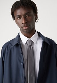 Mornarsko plavi sako preko bijele košulje s svijetlosivim kravatom. Sako ima ovratnik i dugmad, s glatkom teksturom.