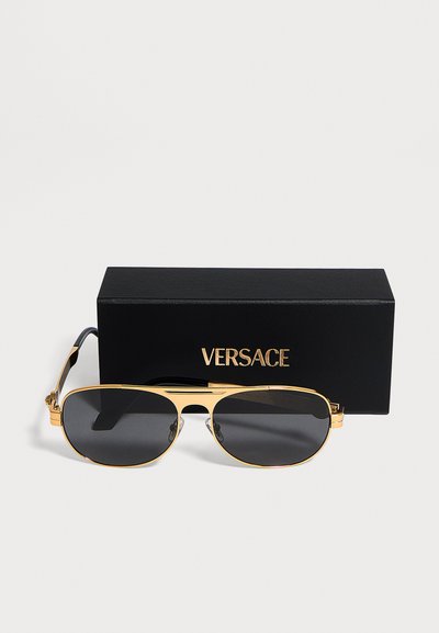 Γυαλιά ηλίου Versace με χρυσή κορνίζα και σκούρους φακούς, τοποθετημένα μπροστά από μια μαύρη θήκη με χρυσό λογότυπο Versace.
