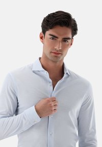 Hellblaues Hemd mit strukturiertem Gewebe, featuring einen spitzen Kragen und langen Ärmeln, teilweise am Hals aufgeknöpft.