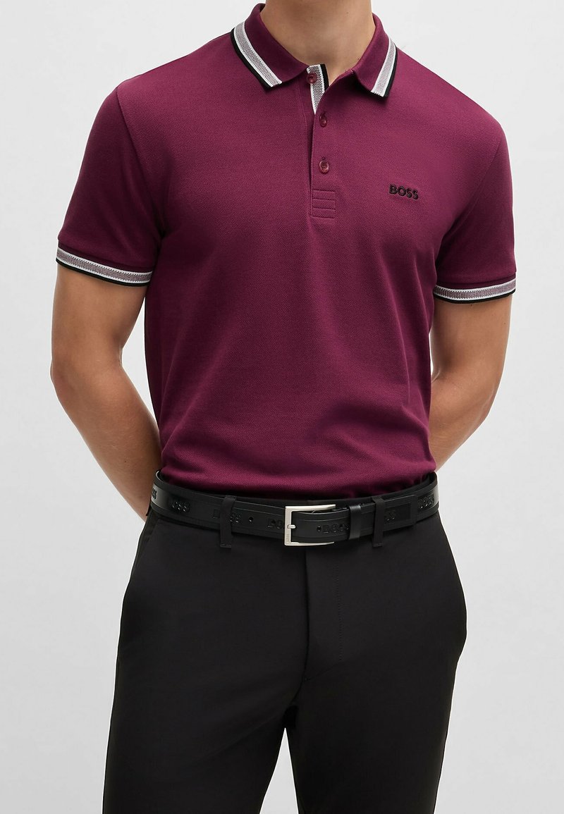 Homme portant un polo violet foncé avec des bordures blanches et noires au col et aux manches, assorti d'un pantalon noir et d'une ceinture noire.