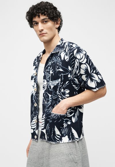 Mlajši moški s kodrastimi lasmi, oblečen v mornarsko modro in belo srajco s kratkimi rokavi z rožnatim vzorcem, preko bele majice brez rokavov in sivimi hlačami.