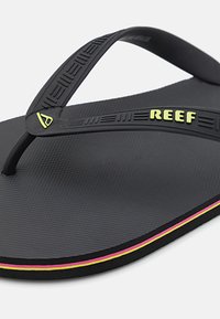 Crne gumene japanke s teksturiranim uloškom. Imaju crni remen s uzdignutim linijama i neon zeleni "REEF" logo, naglašen višebojnim obrubom.