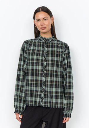 Woman with straight dark hair wears green and black plaid long-sleeve button-up blouse with ruffled cuffs and collar, standing against white background.
