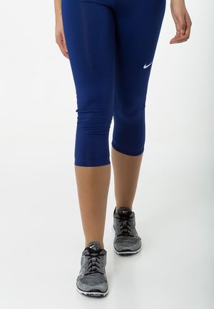 Osoba nosząca niebieskie krótkie legginsy i czarno-szare buty do biegania Nike idąca po białej powierzchni.