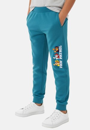 Kind trägt türkisfarbene Jogginghose mit Paw Patrol-Charakteren und Logo am Oberschenkel, kombiniert mit weißen Turnschuhen und einem grauen Shirt.