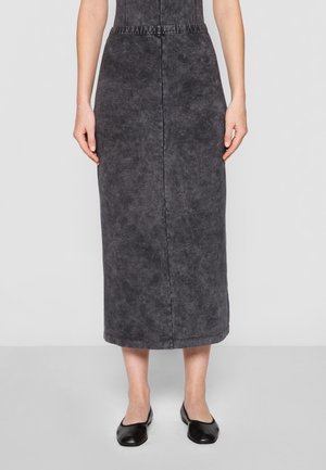 Woman wearing a faded black mid-calf skirt and black flats, standing against a plain light gray background.