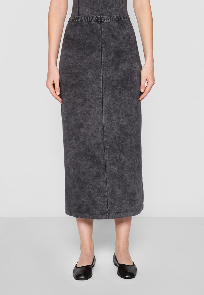 Femme portant une jupe noire délavée mi-mollet et des ballerines noires, debout devant un fond gris clair uni.