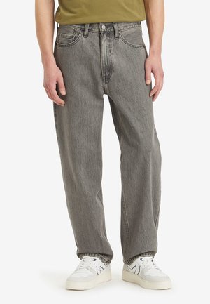 Mann trägt lockere graue Jeans und weiße Turnschuhe, steht vor einem schlichten weißen Hintergrund.