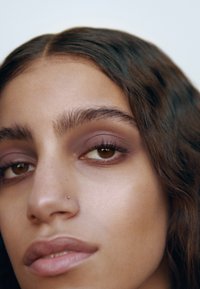Close-up of a face showing smooth skin with a light tan, straight dark hair, and subtle brown eyes. Features include a small nose ring and soft makeup.