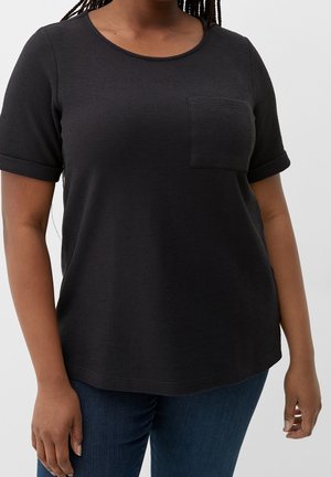 Vrouw draagt een zwart textuur kortemouwshirt met een borstzak, gecombineerd met blauwe jeans, staand tegen een effen achtergrond.