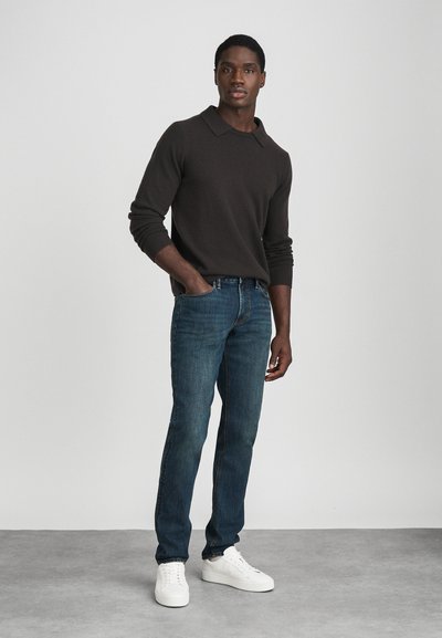 Fiatal férfi, aki egy kézzel a zsebében áll, sötétbarna pulóvert, kék farmernadrágot és fehér sportcipőt visel, sima, világos háttér előtt.