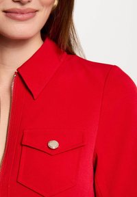 Chaqueta roja con cremallera y cuello puntiagudo, bolsillo en el pecho con un botón a presión plateado y una textura suave.