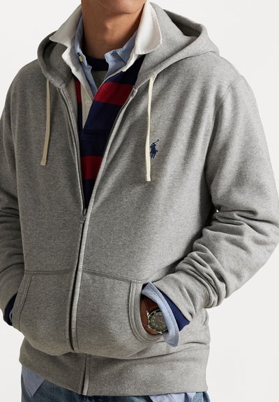 Grå hettegenser med glidelås laget av mykt stoff, med en logo på brystet, snorhette og frontlommer, lagd over en stripete polo.
