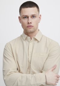 Bež srajca s zaklopom in žepom, izdelana iz teksturiranega blaga. Model ima kratke lase in nosi srebrno ogrlico. Roke prekrižane.