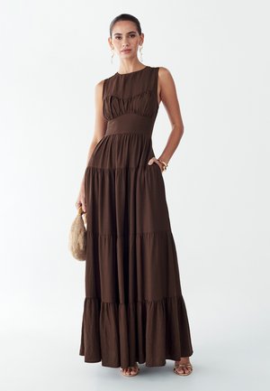 Žena má na sebe hnedé bezrukávové maxi šaty s volánmi, drží tkanú kabelku, doplnená zlatými náušnicami, náramkami a sandálmi.