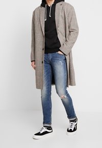 Rutig kappa i ljus beige och svart mönster, lager på lager över en svart hoodie. Bärs med slitna blå jeans och svarta sneakers.