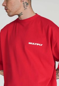 Camiseta de algodón roja de corte holgado con cuello redondo y mangas cortas. Presenta un logotipo impreso en blanco "MULTIPLY" en la parte izquierda del pecho.