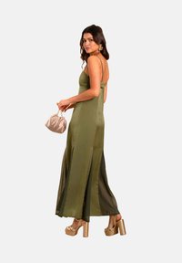 Abito in raso verde oliva con spalline sottili, spacchi laterali e una silhouette fluida. Abbinato a una pochette beige chiaro e a tacchi a piattaforma dorati.