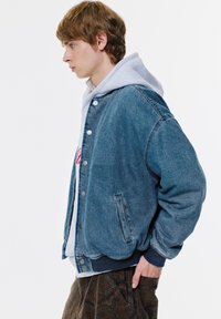 Geacă bomber din denim cu un aspect albastru decolorat, cu butoane tip snap, buzunare laterale și talie elastică. Hanorac gri ușor sub ea.