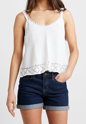 Woman wearing sleeveless white crochet top with scalloped hem and dark blue cuffed denim shorts, hand in pocket, against plain background.