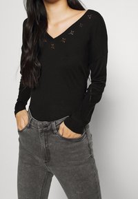 Top negro de manga larga con escote en V y bordado floral alrededor del escote, combinado con jeans gris de cintura alta.
