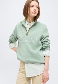 Νεαρό άτομο που φοράει ανοιχτό πράσινο φούτερ Barbour με φερμουάρ μέχρι τη μέση, πάνω από μια ριγωτή πουκάμισα με κουμπιά και μπεζ παντελόνι, κοιτάζοντας προς τα πλάγια.