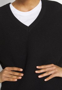 Czarny sweter dziany o fakturze z dekoltem w serek, noszony na białej koszulce, ręce spoczywają na biodrach, prezentując wzór tkaniny.